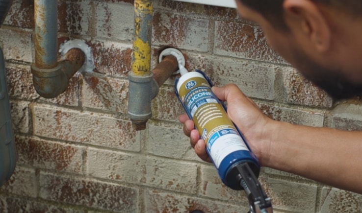 Photo of a person using a caulk gun to apply sealant around pipes against a brick wall.