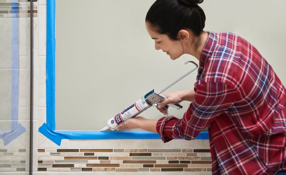 Photo of a woman applying caulk to a tiled bathroom corner with blue painter's tape.