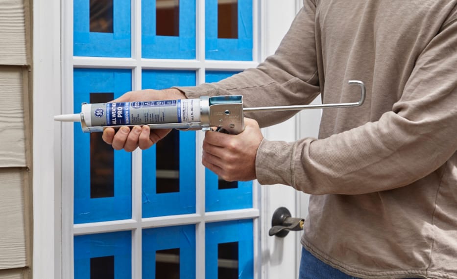 Photo of a person applying GE Pro Silicone sealant to a door frame taped with blue painter's tape.