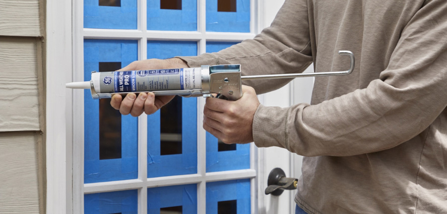 Photo of a person using GE Paintable Acrylic caulk on a window with blue tape for precision.