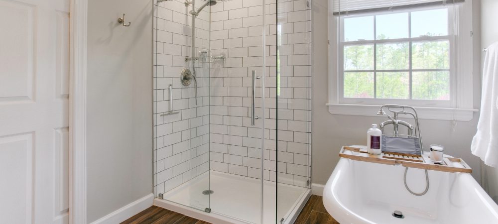 A bright bathroom features a glass-enclosed tile shower and a freestanding tub under soft ceiling and natural window light.