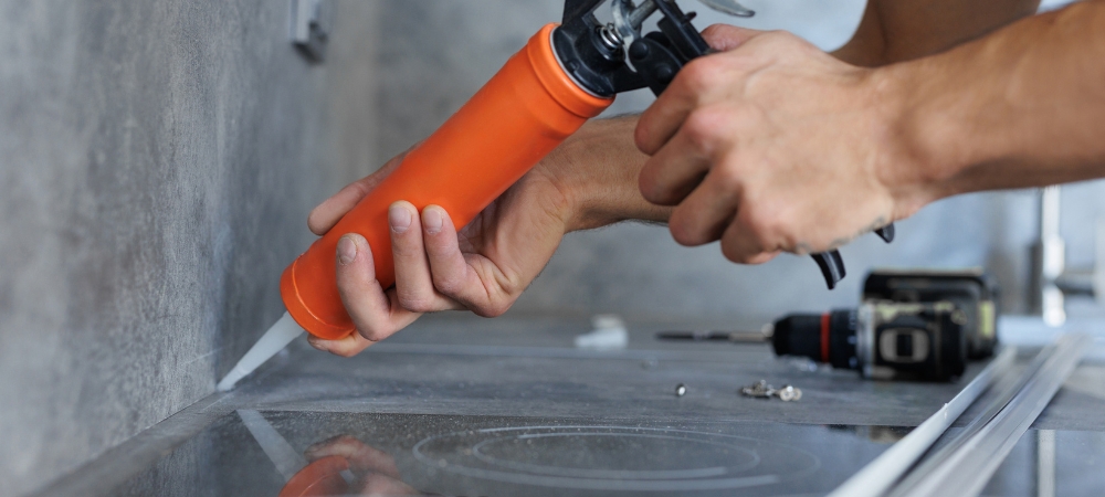 A homeowner applying GE high-temperature silicone sealant behind a kitchen stove to protect against heat and moisture.
