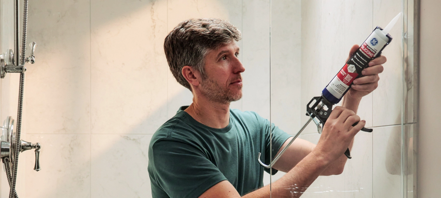 A homeowner applying GE silicone bathroom sealant inside a tiled shower.