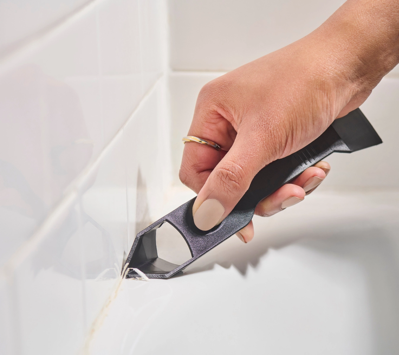 A gloved hand using the GE Sealant Remover Tool to lift a line of old caulk from a bathroom tile joint.