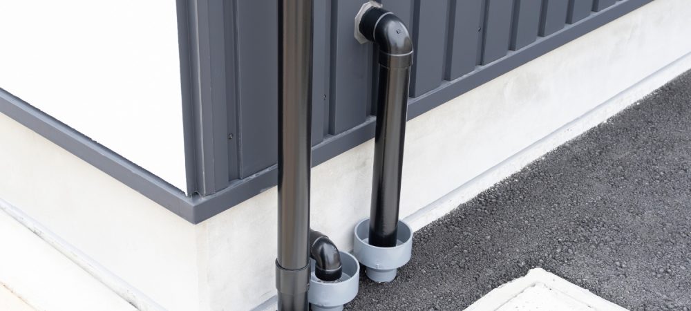 Two black exterior pipes run along the corner of a gray metal-sided building, connecting into the ground through circular fittings in a gravel surface.