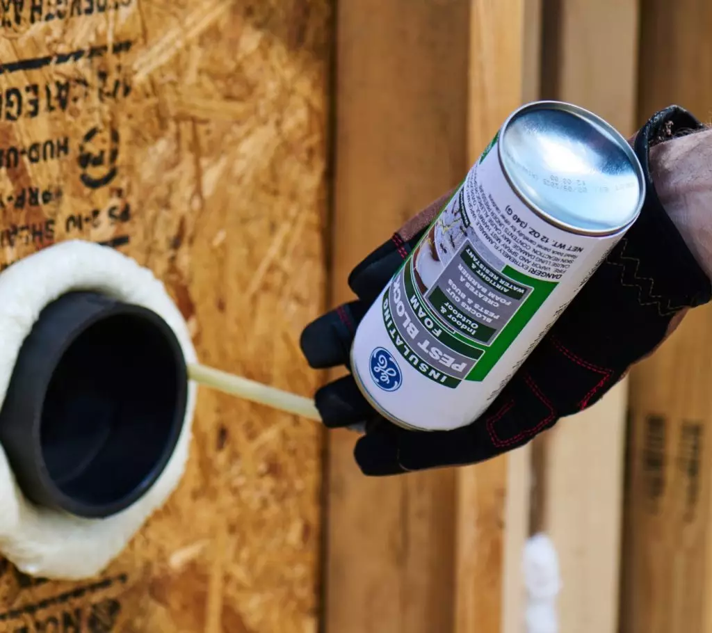 A gloved hand uses GE Pest Block Insulating Foam can to seal around a pipe opening in a wooden wall.
