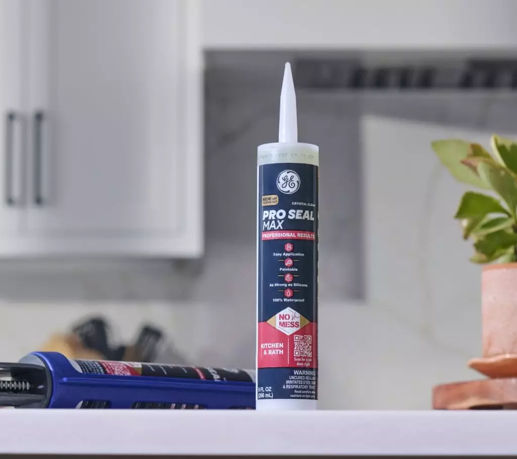 GE Pro Seal Max Kitchen & Bath sealant cartridge stands upright on a kitchen counter next to a caulking gun, with cabinets and a potted plant in the background.