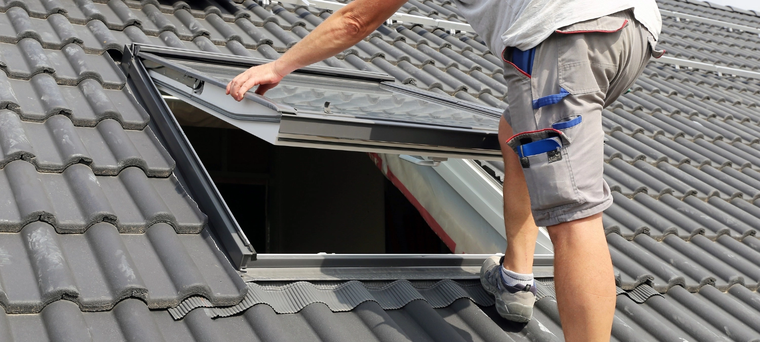 A DIYer applying sealant around a skylight using a caulking gun and safety harness.