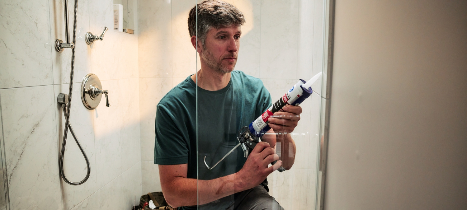 Property manager applying GE 100% Supreme Silicone Kitchen & Bath sealant around a shower to prevent water damage in a rental bathroom.