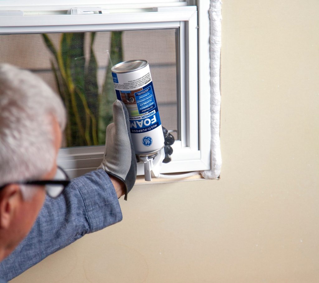 Man using insulating foam to seal around a window frame on the inside of a house.