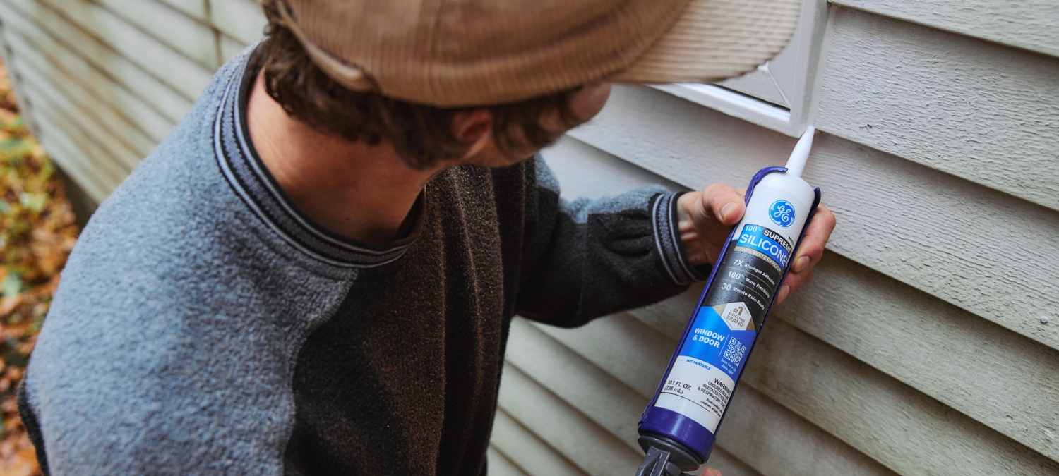 Homeowner sealing a drafty window with GE Supreme Silicone Window & Door silicone sealant to improve energy efficiency and lower heating costs.