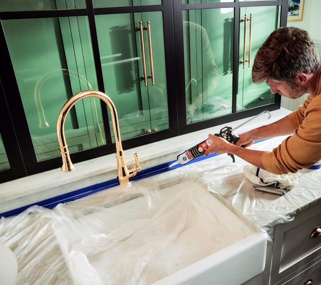 Homeowner applying GE Supreme Silicone Kitchen & Bath Sealant along a kitchen sink to protect wet areas from leaks as part of their annual home maintenance checklist.