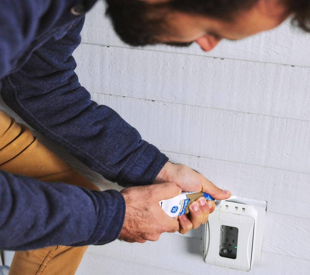 Man is applying GE Advanced Silicone Window & Door Sealant around an exterior electrical outlet.