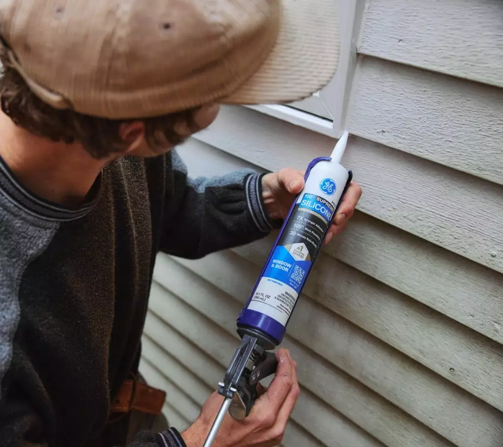 A photo of a homeowner applying sealant around an exterior window frame.