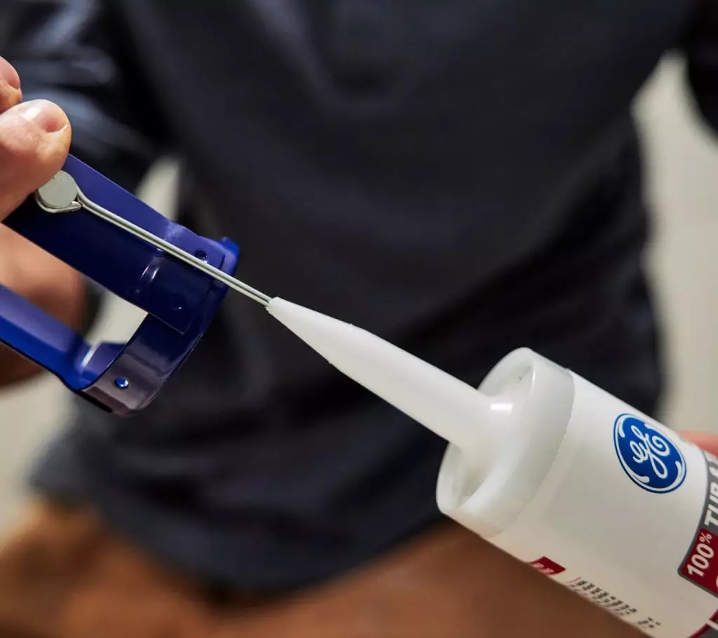 A photo of a homeowner using a nail to break the inner seal on a tube of caulk.