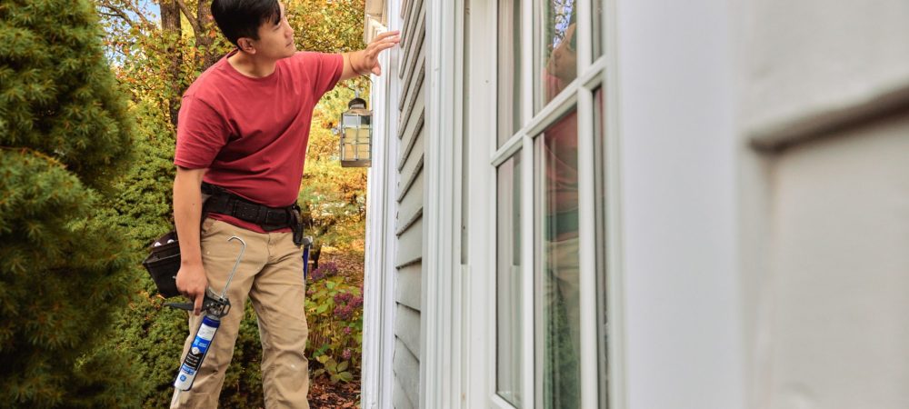 A contractor inspecting the exterior of a house while holding a caulking gun.